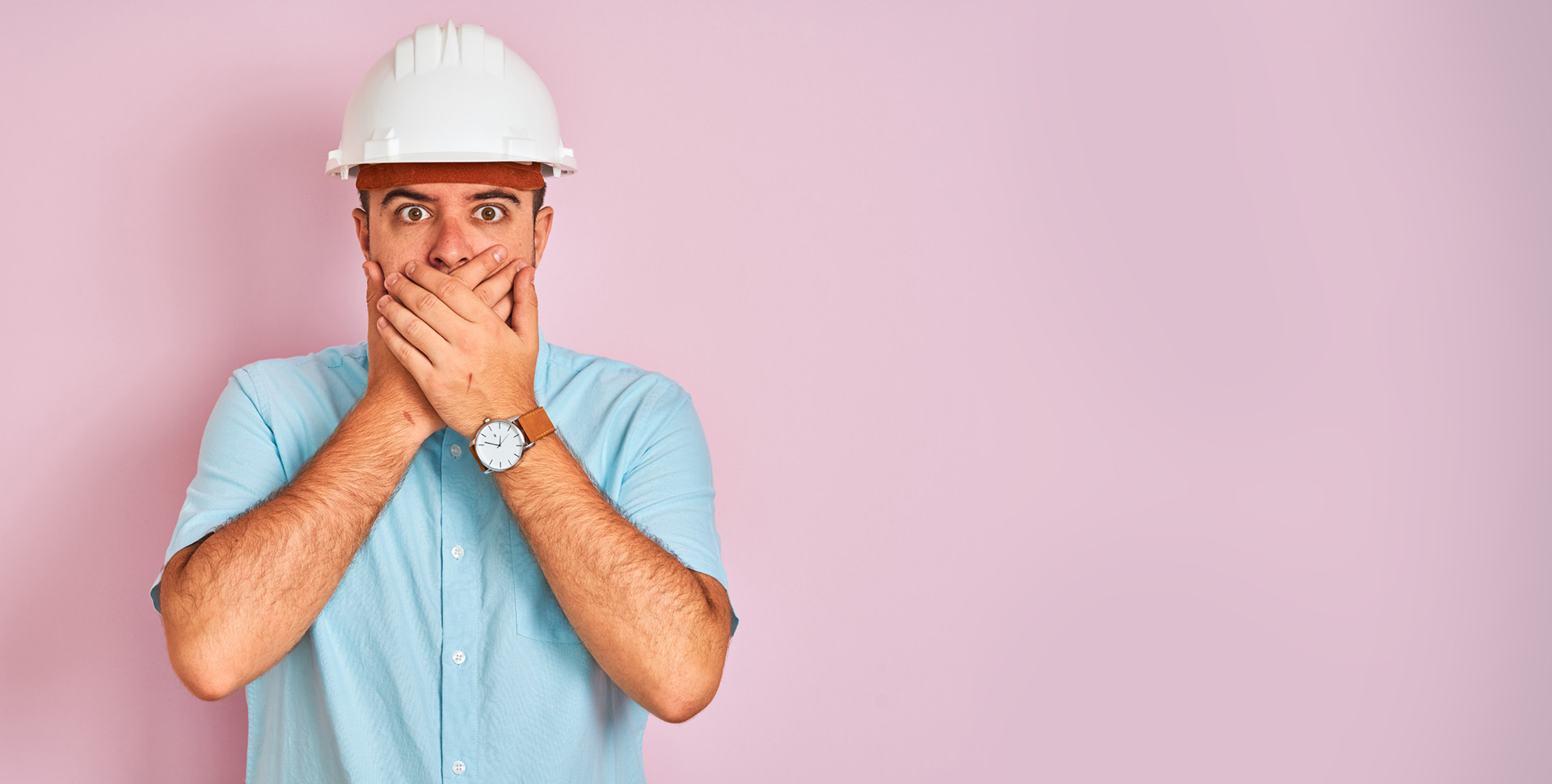 An image of a male worker with a light blue shirt and white hardhat standing in front of a light pink background. He is covering is mouth with both hands.