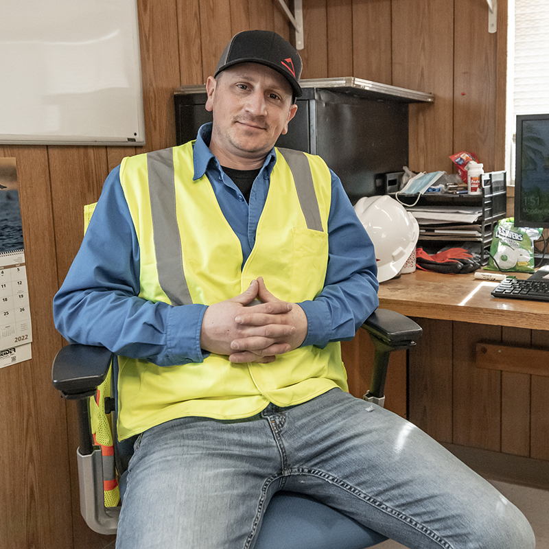 An image of a male employee wearing a safety vest and baseball hat. He sits in an office chair hands interlaced facing the camera.